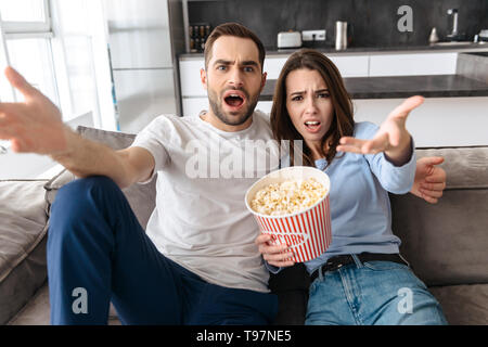 Immagine di orinato giovane uomo e donna in abbigliamento casual mangiare popcorn dalla benna mentre è seduto sul lettino piscina e guardare film Foto Stock