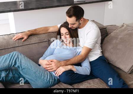 Immagine del Caucaso la donna incinta e uomo bello in abbigliamento casual abbracciando insieme a casa mentre sdraiato sul divano nel soggiorno Foto Stock