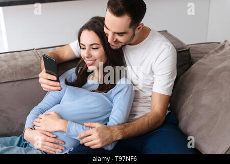 Immagine positiva della donna incinta e uomo bello in abbigliamento casual sdraiato sul divano nel soggiorno di casa e utilizza lo smartphone Foto Stock