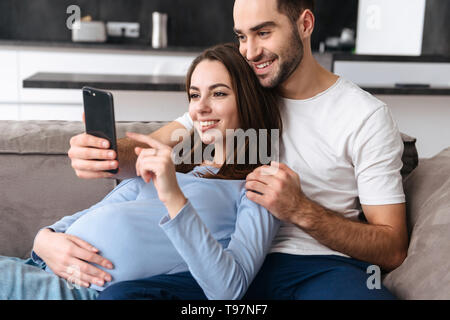 Immagine della comunità donna incinta e uomo bello in abbigliamento casual sdraiato sul divano nel soggiorno di casa e utilizza lo smartphone Foto Stock