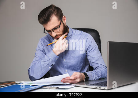 Concentrato barbuto uomo elegante in occhiali leggendo qualcosa mentre seduti a tavola in office Foto Stock