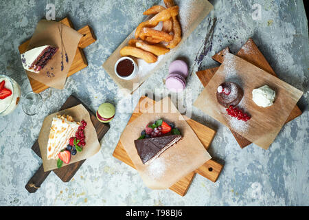 Un assortimento di dolci: torta, churos, rotolo di meringa, gelato, decorata con frutti di bosco su tavole di legno. Vista dall'alto. Grigio sfondo a trama. Bellissimi piatti di servizio. Il dessert. La catena alimentare Foto Stock