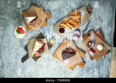 Un assortimento di dolci: torta, churos, rotolo di meringa, gelato, decorata con frutti di bosco su tavole di legno. Vista dall'alto. Grigio sfondo a trama. Bellissimi piatti di servizio. Il dessert. La catena alimentare Foto Stock