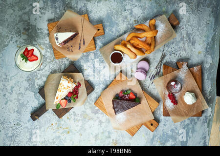 Un assortimento di dolci: torta, churos, rotolo di meringa, gelato, decorata con frutti di bosco su tavole di legno. Vista dall'alto. Grigio sfondo a trama. Bellissimi piatti di servizio. Il dessert. La catena alimentare Foto Stock