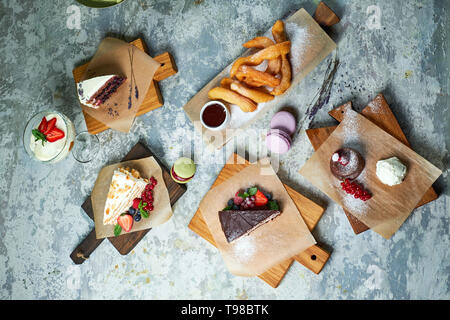 Un assortimento di dolci: torta, churos, rotolo di meringa, gelato, decorata con frutti di bosco su tavole di legno. Vista dall'alto. Grigio sfondo a trama. Bellissimi piatti di servizio. Il dessert. La catena alimentare Foto Stock