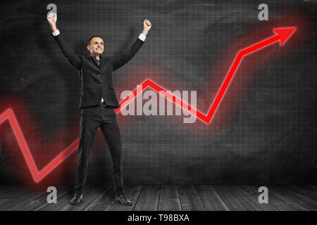 Parte anteriore a lunghezza piena vista di happy businessman bracci di sollevamento nel gesto trionfante con il grafico rosso freccia salendo dietro di lui vicino al nero grafico governata da parete. Foto Stock