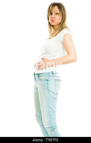 Blonde slim donna in maglia bianca e jeans mostra espressioni di obesità e sovrappeso - sbuffando le guance e grande stomaco - sfondo bianco Foto Stock