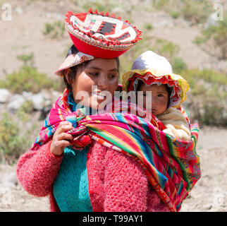 Sorridente Quechua Donna vestita in colorate a mano tradizionale vestito e portando il suo bambino In una fionda. Ottobre 21, 2012 - Patachancha, Cuzco, Perù Foto Stock