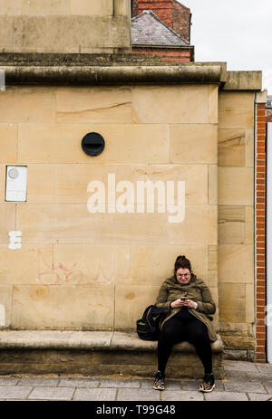 Donna seduta su un banco di lavoro, appoggiata contro la parete utilizzando lo smartphone, Quayside, Newcastle upon Tyne, England, Regno Unito Foto Stock