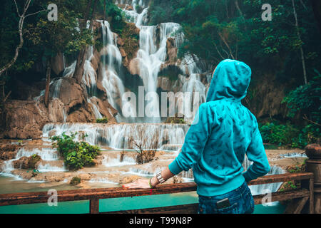 Blonde Backpacker o Traveler Ragazza in un abito blu guardando sopra le cascate di Kuang Si in Laos. Una giovane donna guarda e si ammira la splendida cascata in Foto Stock