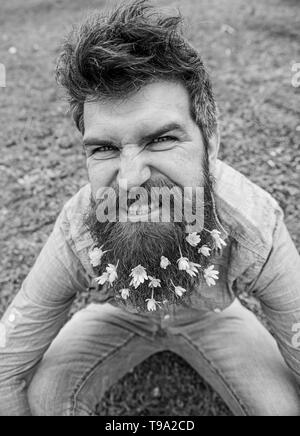 Ragazzo con minor celandine fiori in barba tenendo selfie foto. Hipster con arrabbiato grimace sulla faccia tenendo selfie foto. Mad concetto dell'umore. L'uomo gode di molla, prato verde, sfondo sfocato. Foto Stock