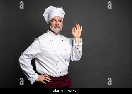 Lo Chef rendere gustoso gesto delizioso da baciare le dita. Fiducioso barbuto chef maschio in bianco uniforme con segno perfetto. Foto Stock