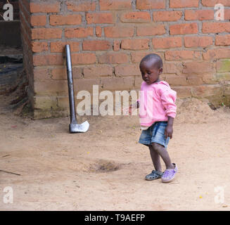 Un bambino del Malawi cammina davanti a una grande ascia metallica sinistra appoggiata contro un muro dopo la trinciatura di legna da ardere Foto Stock