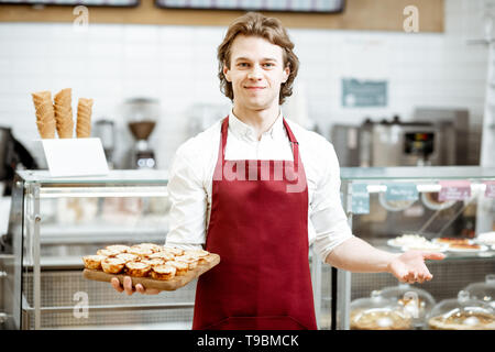 Ritratto di un bel venditore o pasticcere in rosso grembiule in piedi con la fresca di forno pastel de nata nella pasticceria Foto Stock