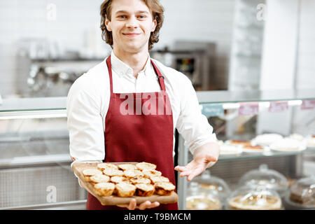 Ritratto di un bel venditore o pasticcere in rosso grembiule in piedi con la fresca di forno pastel de nata nella pasticceria Foto Stock