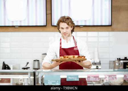 Ritratto di un bel venditore o pasticcere in rosso grembiule in piedi con la fresca di forno pastel de nata nella pasticceria Foto Stock