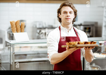 Ritratto di un bel venditore o pasticcere in rosso grembiule in piedi con la fresca di forno pastel de nata nella pasticceria Foto Stock