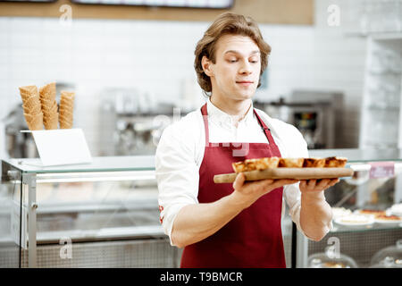 Ritratto di un bel venditore o pasticcere in rosso grembiule in piedi con la fresca di forno pastel de nata nella pasticceria Foto Stock