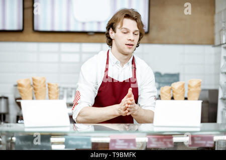 Bello venditore tenendo cono di cialda durante la preparazione del gelato per il client nella moderna pasticceria Foto Stock