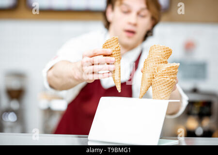 Bello venditore tenendo cono di cialda durante la preparazione del gelato per il client nella moderna pasticceria Foto Stock