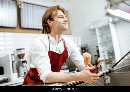 Bello venditore nel grembiule rosso vendita di gelato nella moderna pasticceria Foto Stock