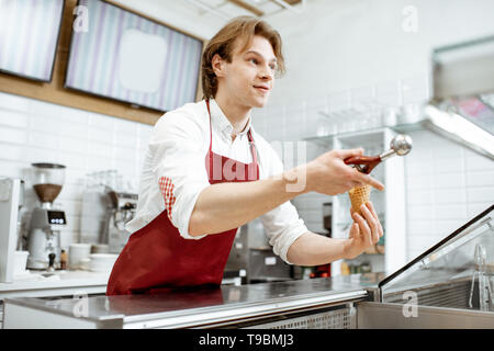 Bello venditore nel grembiule rosso vendita di gelato nella moderna pasticceria Foto Stock