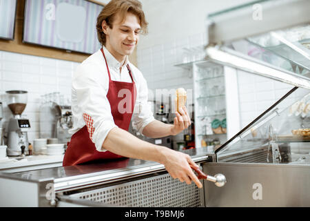 Bello venditore nel grembiule rosso vendita di gelato nella moderna pasticceria Foto Stock