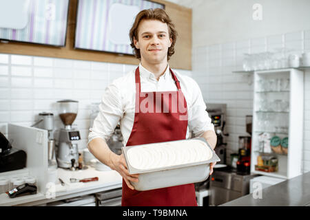 Ritratto di un bel venditore o pasticceria a grembiule rosso tenendo il vassoio con gelato nel negozio Foto Stock