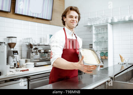 Ritratto di un bel venditore o pasticceria a grembiule rosso tenendo il vassoio con gelato nel negozio Foto Stock