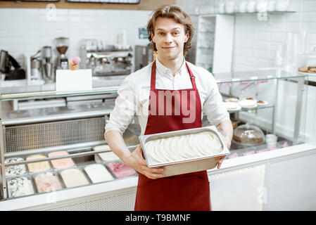 Ritratto di un bel venditore o pasticceria a grembiule rosso tenendo il vassoio con gelato nel negozio Foto Stock