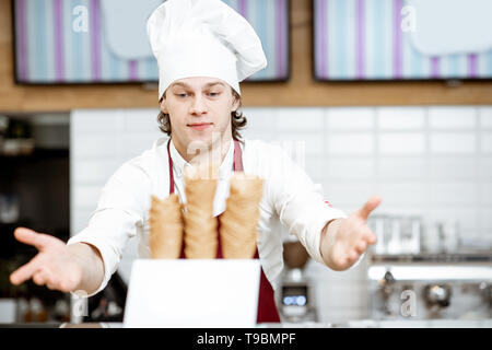 Bello venditore in grembiule e hat prendendo i coni di cialda durante la preparazione del gelato presso la moderna pasticceria Foto Stock