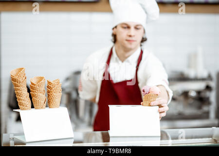 Il giovane chef cuoco o venditore in grembiule e cappello la vendita di gelati in cono di cialda nella moderna pasticceria Foto Stock