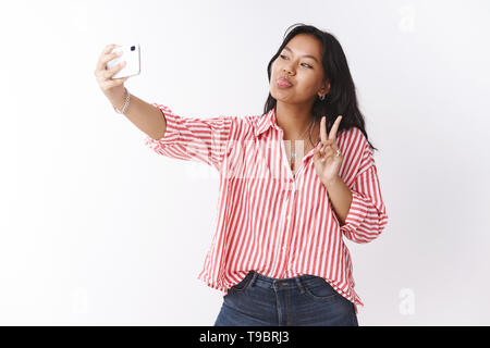 Studio shot di fiduciosa bella giovane ed elegante donna in uscita in camicia a righe scimmiottare e facendo facce buffe sulla fotocamera come prendendo selfie con Foto Stock