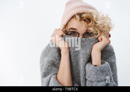 Non è possibile arrivare a me l'inverno. Carino sciocco e giocoso ragazza bionda in bicchieri e beanie tirando il collare del maglione sulla faccia per nascondere e sbirciando fotocamera Foto Stock