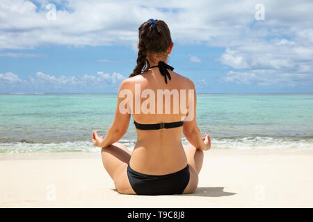 Vista posteriore di una donna in Bikini nero meditando sulla spiaggia vicino al mare turchese Foto Stock