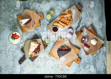 Un assortimento di dolci: torta, churos, rotolo di meringa, gelato, decorata con frutti di bosco su tavole di legno. Vista dall'alto. Grigio sfondo a trama. Bellissimi piatti di servizio. Il dessert. La catena alimentare Foto Stock