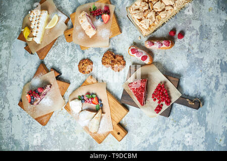 Un assortimento di dolci: torta, churos, rotolo di meringa, gelato, decorata con frutti di bosco su tavole di legno. Vista dall'alto. Grigio sfondo a trama. Bellissimi piatti di servizio. Il dessert. La catena alimentare Foto Stock