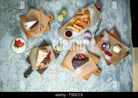 Un assortimento di dolci: torta, churos, rotolo di meringa, gelato, decorata con frutti di bosco su tavole di legno. Vista dall'alto. Grigio sfondo a trama. Bellissimi piatti di servizio. Il dessert. La catena alimentare Foto Stock