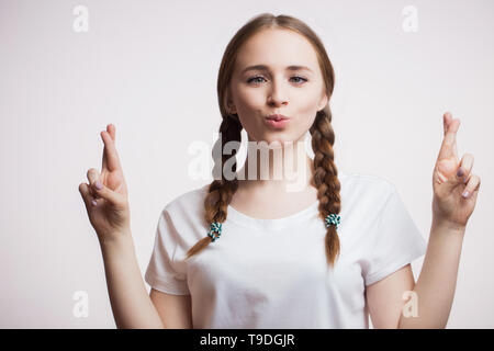 Allegro felice giovane donna con gli occhiali e una t-shirt fa un divertente grimace, incrocia le dita su sfondo bianco. Le emozioni umane, facial expres Foto Stock