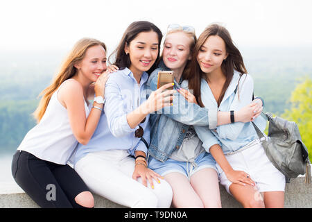 Quattro bellissime donne giovani utilizza lo smartphone e sorridente nel parco, all'aperto. Allegro studente ragazze su sfondo natura Foto Stock