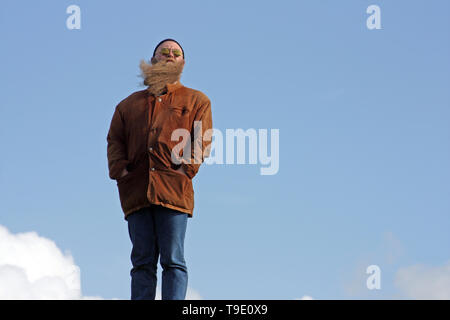 Mann mit langem Bart, der vom Wind verwirbelt wurde, steht am Ufer des Meeres Foto Stock