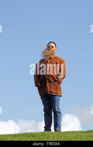 Mann mit langem Bart, der vom Wind verwirbelt wurde, steht am Ufer des Meeres Foto Stock