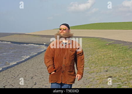 Mann mit langem Bart, der vom Wind verwirbelt wurde, steht am Ufer des Meeres Foto Stock