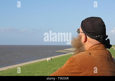 Mann mit langem Bart, der vom Wind verwirbelt wurde, steht am Ufer des Meeres Foto Stock