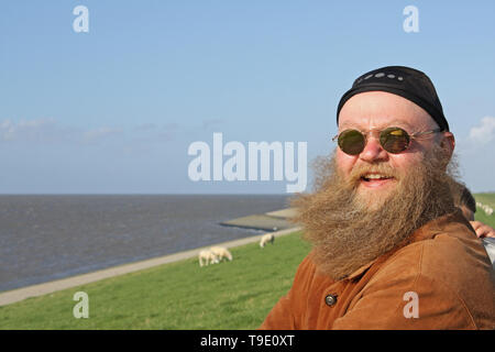 Mann mit langem Bart, der vom Wind verwirbelt wurde, steht am Ufer des Meeres Foto Stock