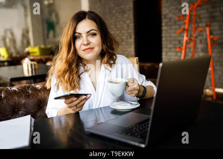 Una giovane donna simpatico e non un thin-intitolata body building, si trova in un accogliente caffetteria, funziona su un computer e può contenere uno smartphone nelle sue mani. Foto Stock