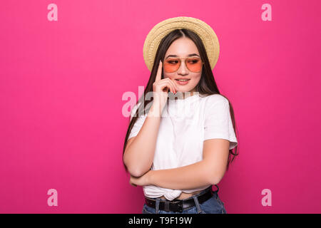 Lieta brunette donna nel cappello di paglia e occhiali da sole in posa mentre guarda lontano su sfondo rosa Foto Stock