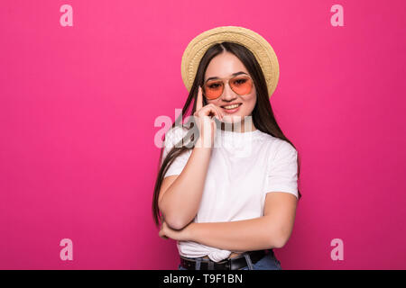 Lieta brunette donna nel cappello di paglia e occhiali da sole in posa mentre guarda lontano su sfondo rosa Foto Stock