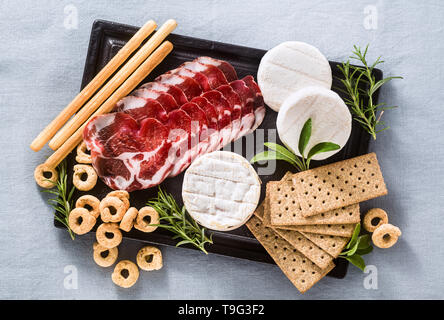 Salumi e formaggi sono serviti su un vassoio su un tavolo con il vino bianco, crackers, grissini e taralli con erbe aromatiche su un lino blu tavola festiva Foto Stock