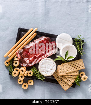 Salumi e formaggi sono serviti su un vassoio su un tavolo con il vino bianco, crackers, grissini e taralli con erbe aromatiche su un lino blu tavola festiva Foto Stock